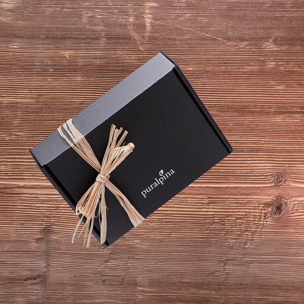 Puralpina Überraschungsbox in schwarzer Geschenkbox mit Bastschleife auf Holzuntergrund