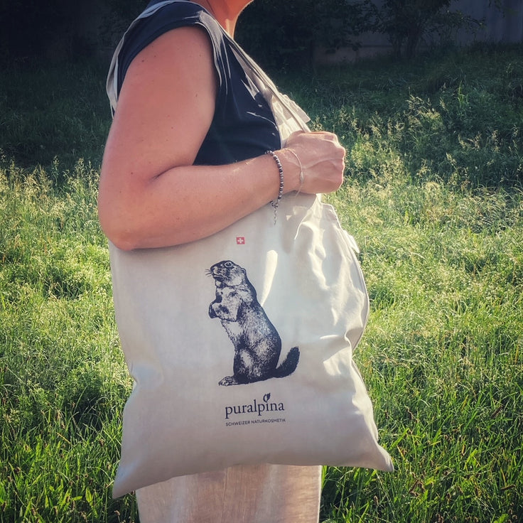 Person carrying a light Puralpina cotton bag with a marmot motif and logo in a green setting.